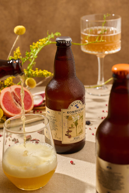 Botella de Kombucha Biloba Elixir sirviendo kombucha en un vaso sobre mesa con ingredientes botánicos.